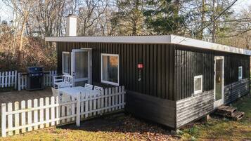 A small, single-story cabin with a white picket fence, a grill, and a door with a flag.