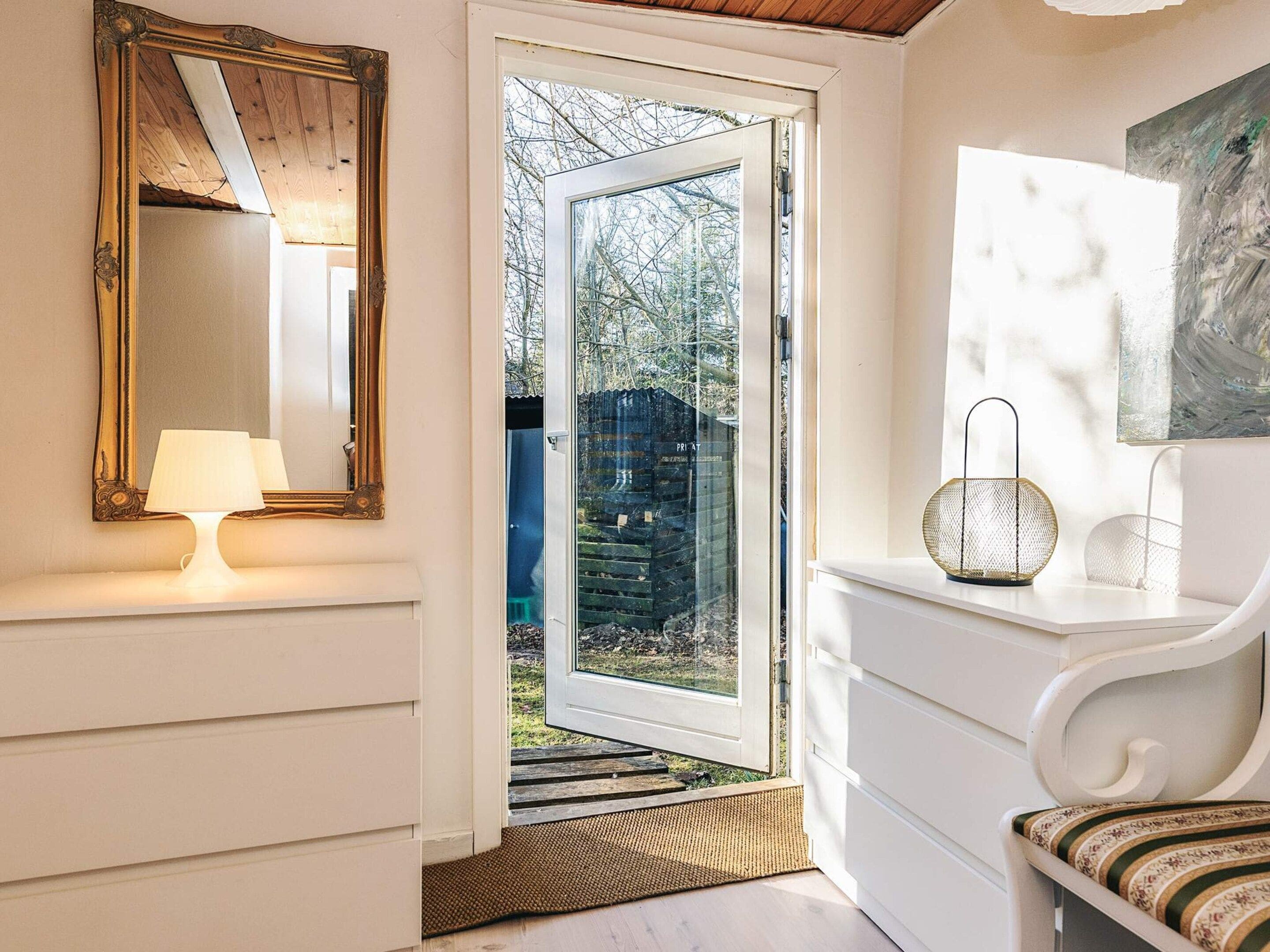 A room with a large mirror, a white chair, a small table with a lamp, and a door leading outside.