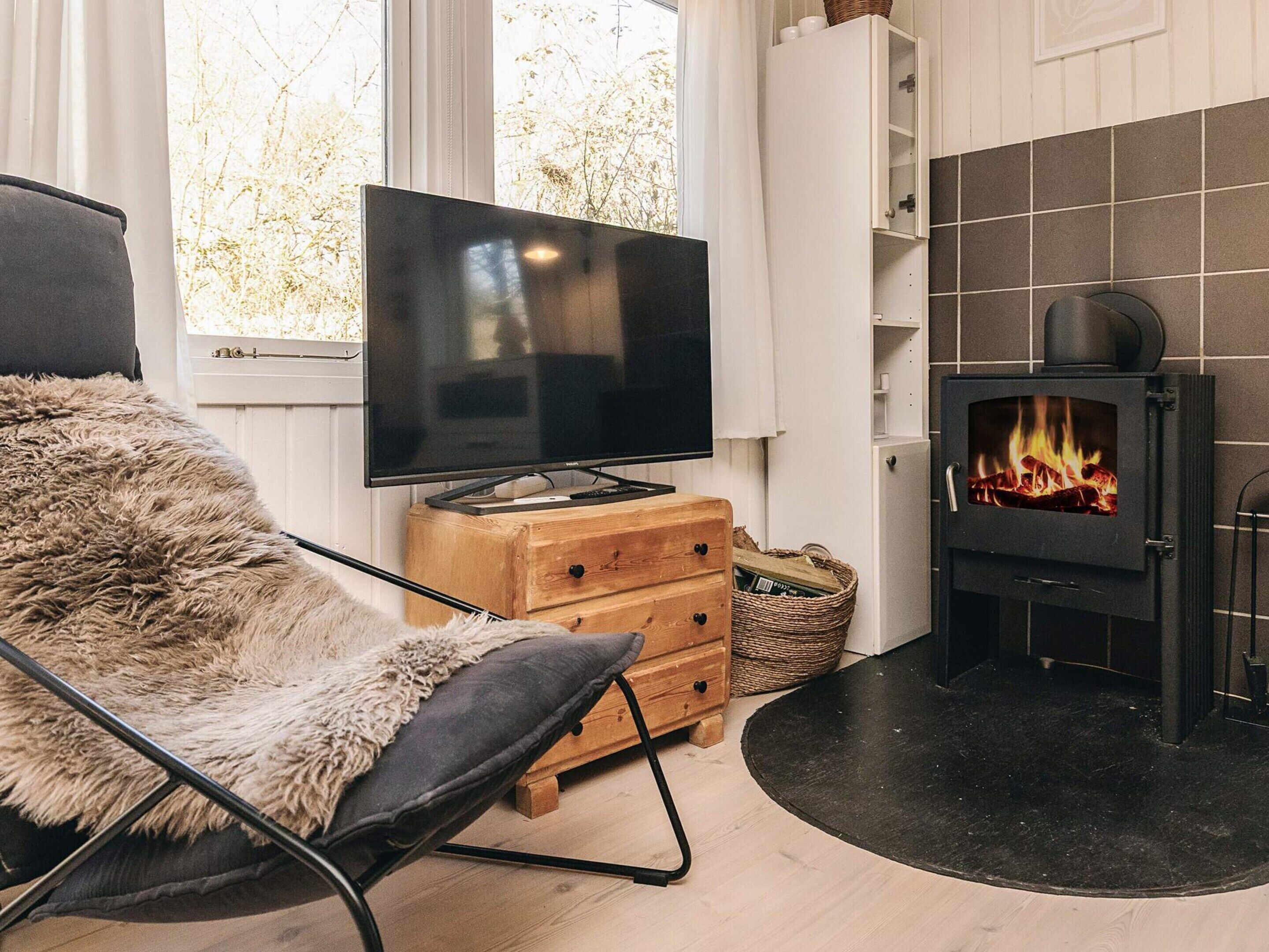 A cozy living room with a flat-screen TV, a fluffy chair, a wooden chest of drawers, and a wood-burning stove.