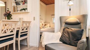 Una habitación acogedora con una silla de madera, un sillón con cojines, una mesita con un florero y una estantería con diversos objetos.