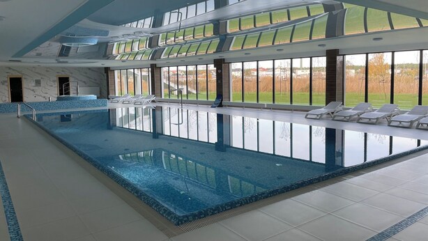 Una amplia piscina cubierta con paredes de vidrio transparente, suelo de baldosas y varias sillas de descanso a orillas de la piscina.