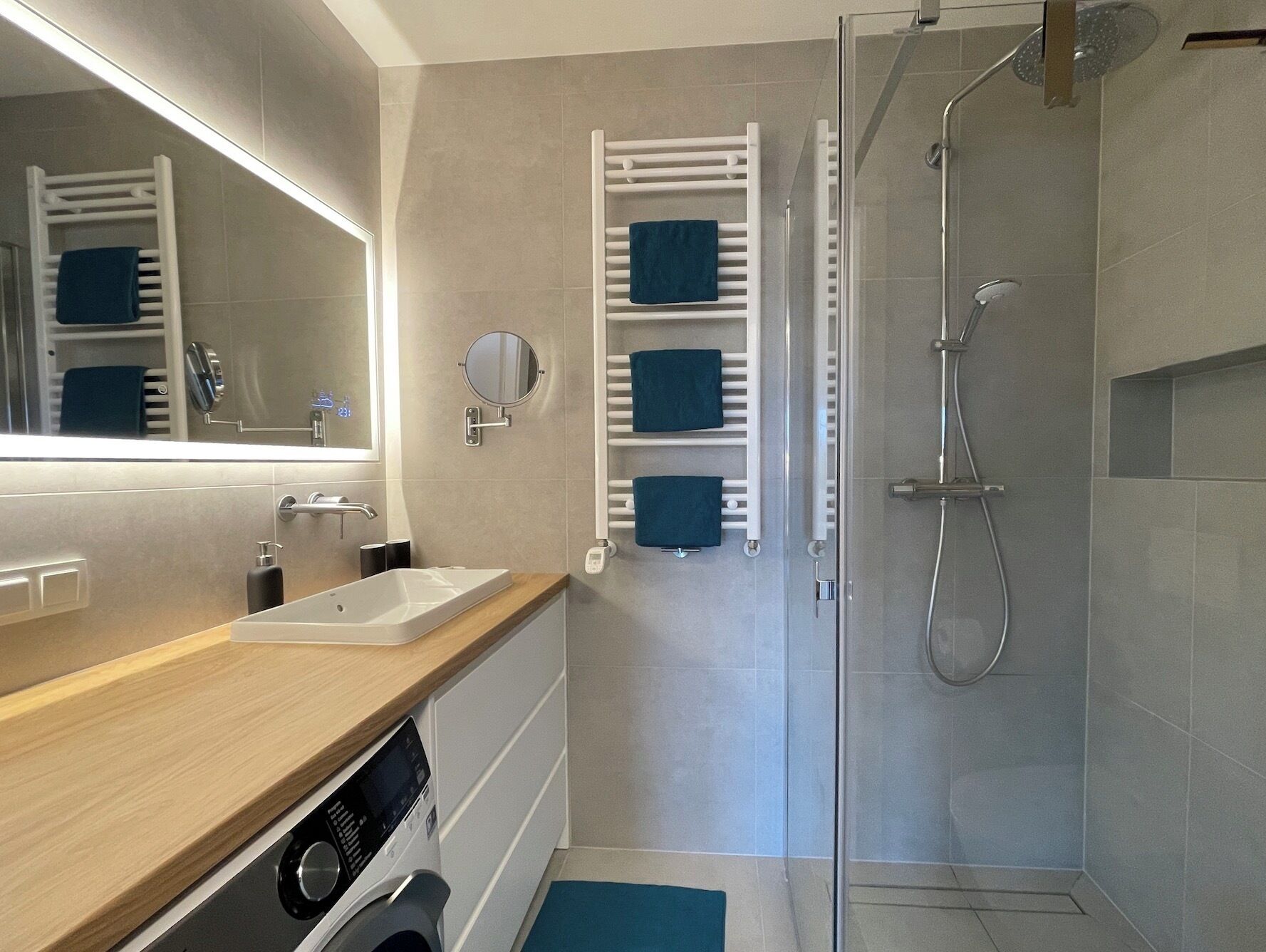 A modern bathroom with a wooden vanity, a built-in washing machine, a glass shower enclosure, and a towel rack with multiple towels.