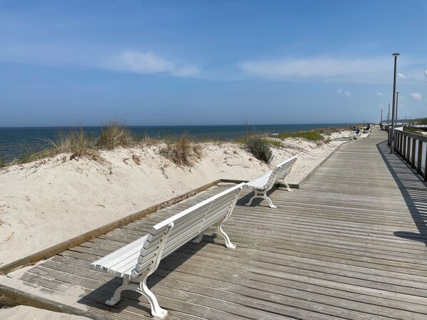 A wooden boardwalk with benches along a sandy beach, leading to the ocean.