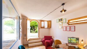 Quarto com escrivaninha, computador, cadeiras e vista para um jardim através de uma porta aberta.