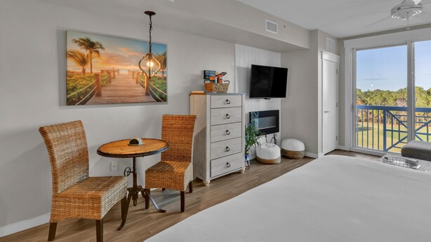 A hotel room with a large bed, a dining table with chairs, a TV, a painting, and a balcony with a view. - Serene 4th Floor Elation Condo: Golf Course and Foliage Views! King Bed, Sleeper Sofa, Kitchenette for Your Convenience at Sandestin Golf and Beach Resort!! (Miramar Beach)
