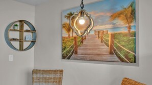 A room with a wooden floor, a round table with a blue fish ornament, wicker chairs, a wall-mounted shelf with decorative items, and a painting of a wooden pier leading to a beach. - Serene 4th Floor Elation Condo: Golf Course and Foliage Views! King Bed, Sleeper Sofa, Kitchenette for Your Convenience at Sandestin Golf and Beach Resort!! (Miramar Beach)
