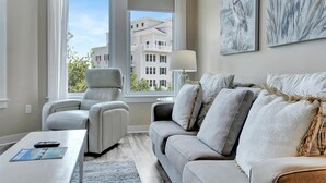 A modern living room with a grey sofa, a white armchair, and a coffee table. There is a window with a view of buildings outside.