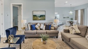 A living room with a beige sofa, a blue armchair, and a coffee table with a plant. There is a painting on the wall and a chandelier. - Spacious 3-Bedroom Retreat with Lake Views (Miramar Beach)