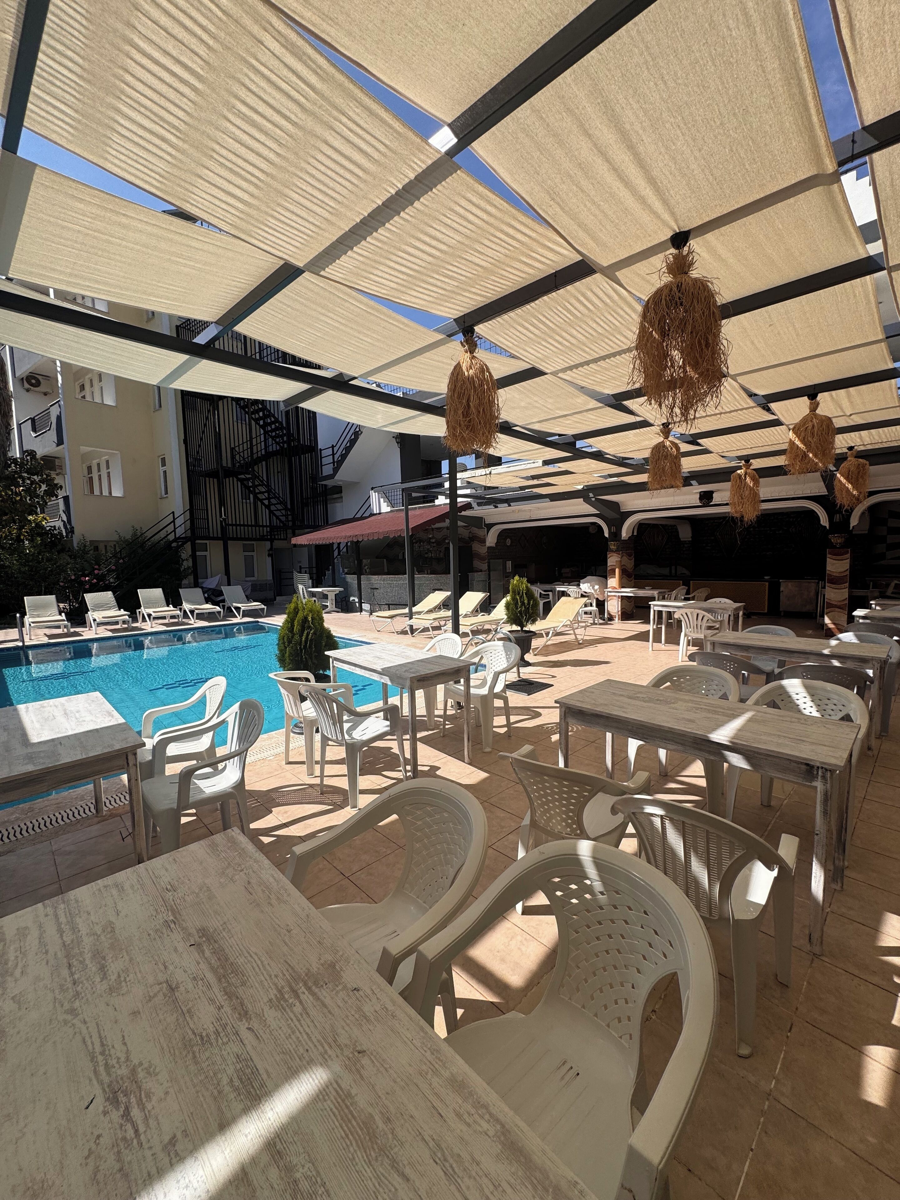 A poolside area with lounge chairs and tables, a swimming pool, and a covered seating area with hanging decorations.