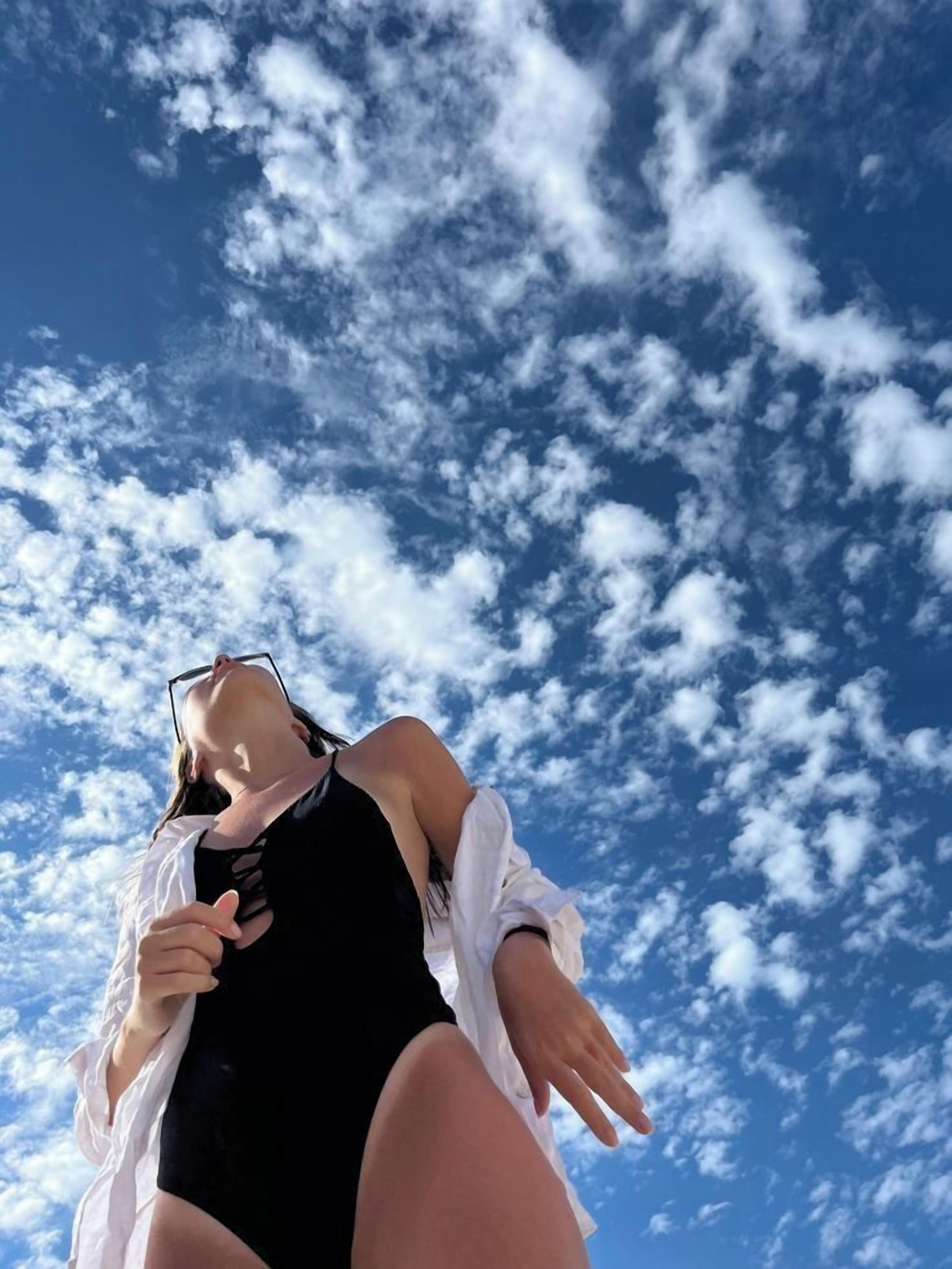 Une personne vêtue d’un maillot de bain noir et d’une chemise blanche, se tenant devant un ciel nuageux.