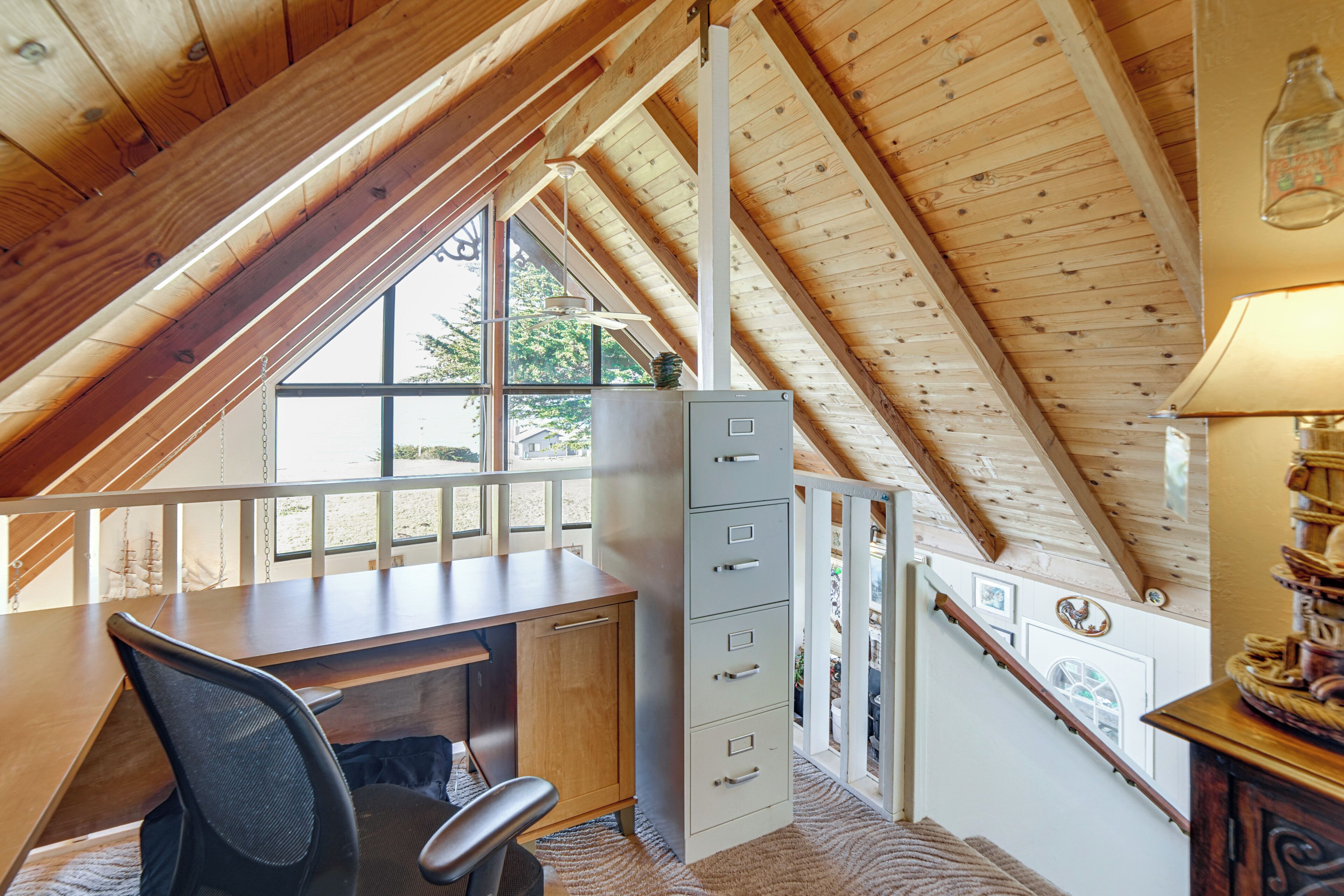 A loft bedroom with a sloped ceiling, a desk, a chair, a filing cabinet, and a window with a view of trees.