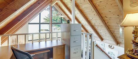 A loft bedroom with a sloped ceiling, a desk, a chair, a filing cabinet, and a window with a view of trees.