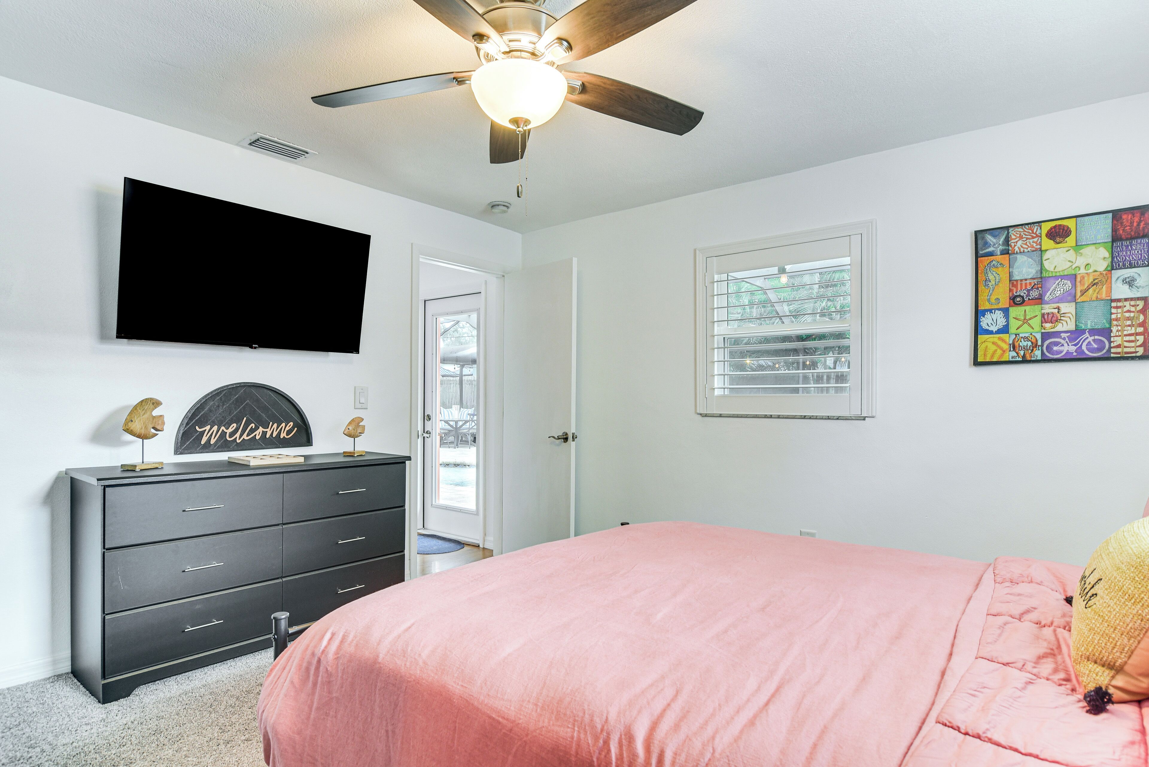 A bedroom with a ceiling fan, a TV mounted on the wall, a dresser, a bed with a pink bedspread, and a window with blinds.