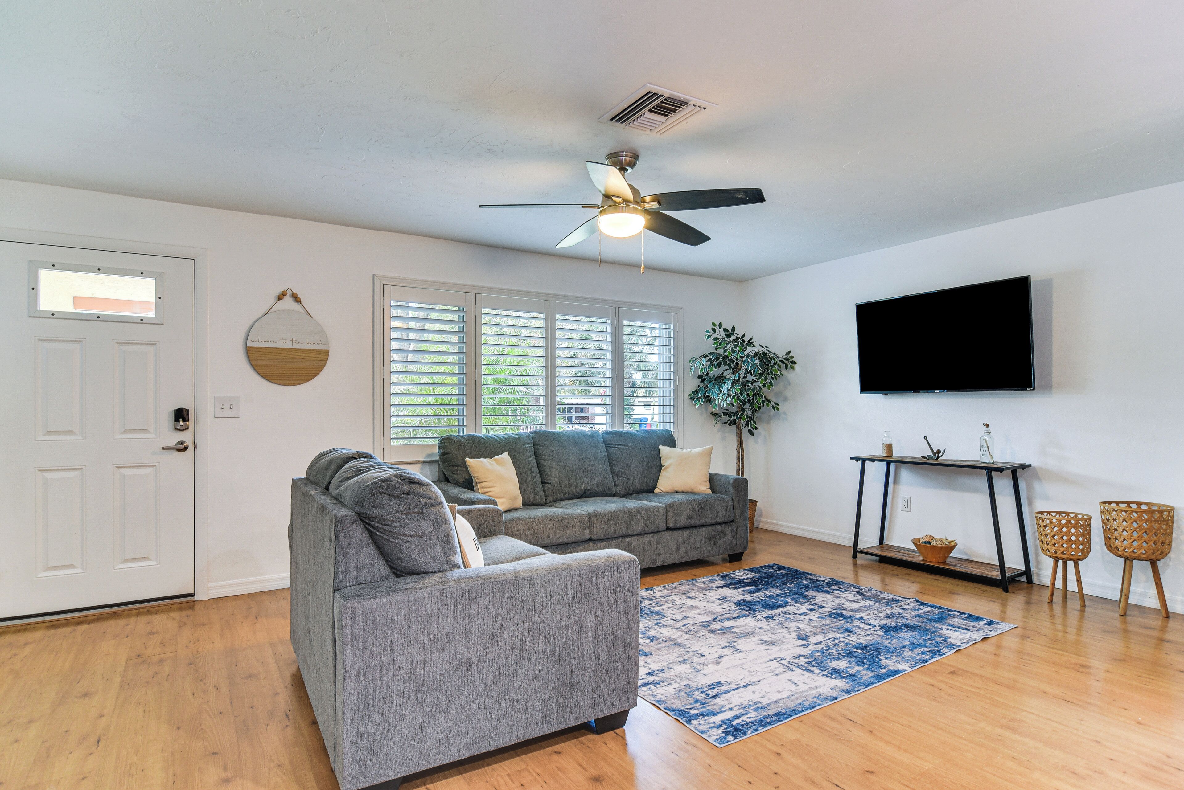 A living room with two grey sofas, a ceiling fan, a large window with blinds, a flat-screen TV, and a small table with chairs.