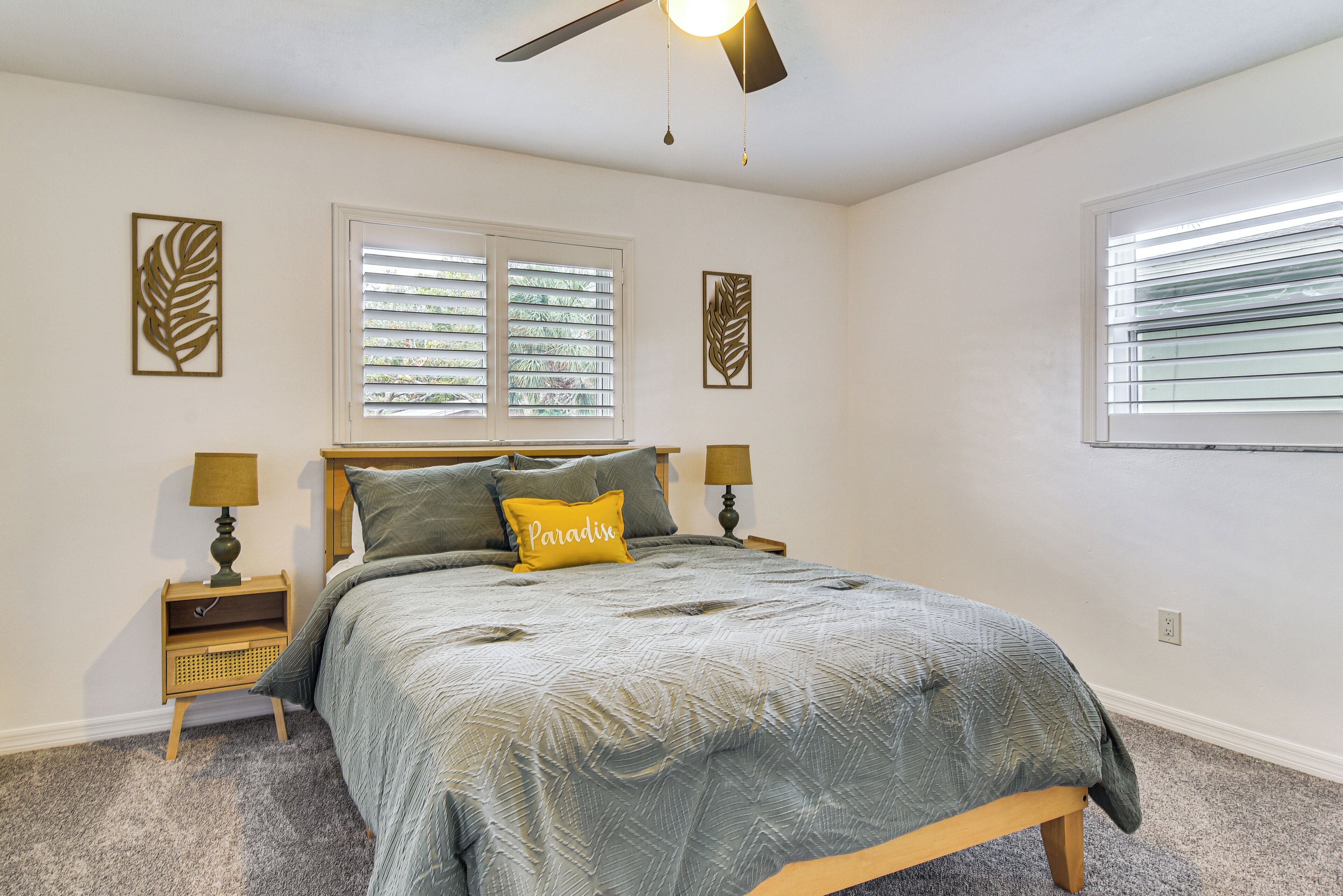 A bedroom with a bed, two nightstands, a ceiling fan, and two windows with blinds.