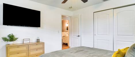 A bedroom with a bed, a ceiling fan, a TV, a dresser, and a door leading to a bathroom.