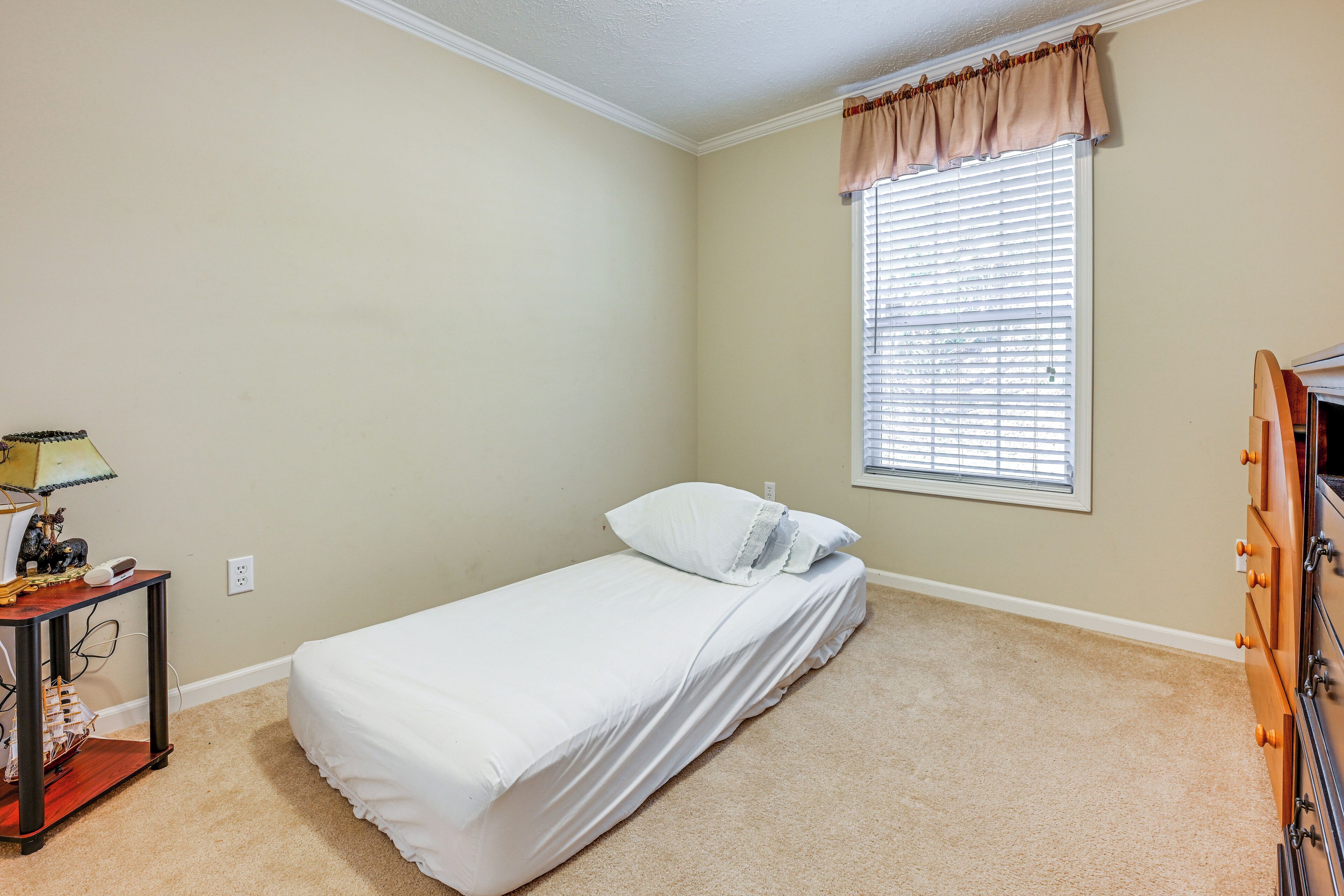 A small, neatly arranged bedroom with a single bed, a nightstand with a lamp, and a dresser.