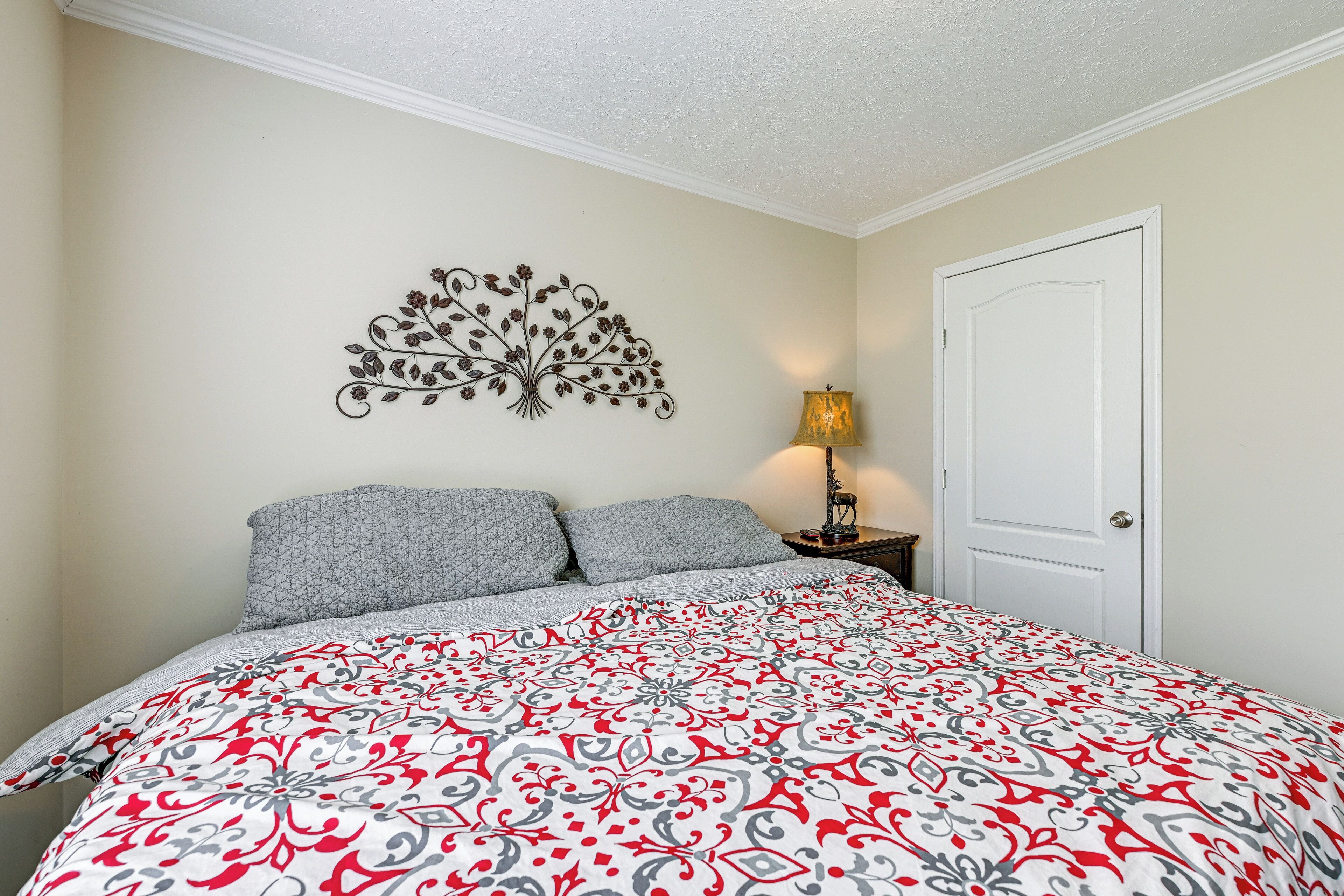 Un dormitorio con cama, dos almohadas, un adorno en la pared, una mesita de noche con lámpara y una puerta.