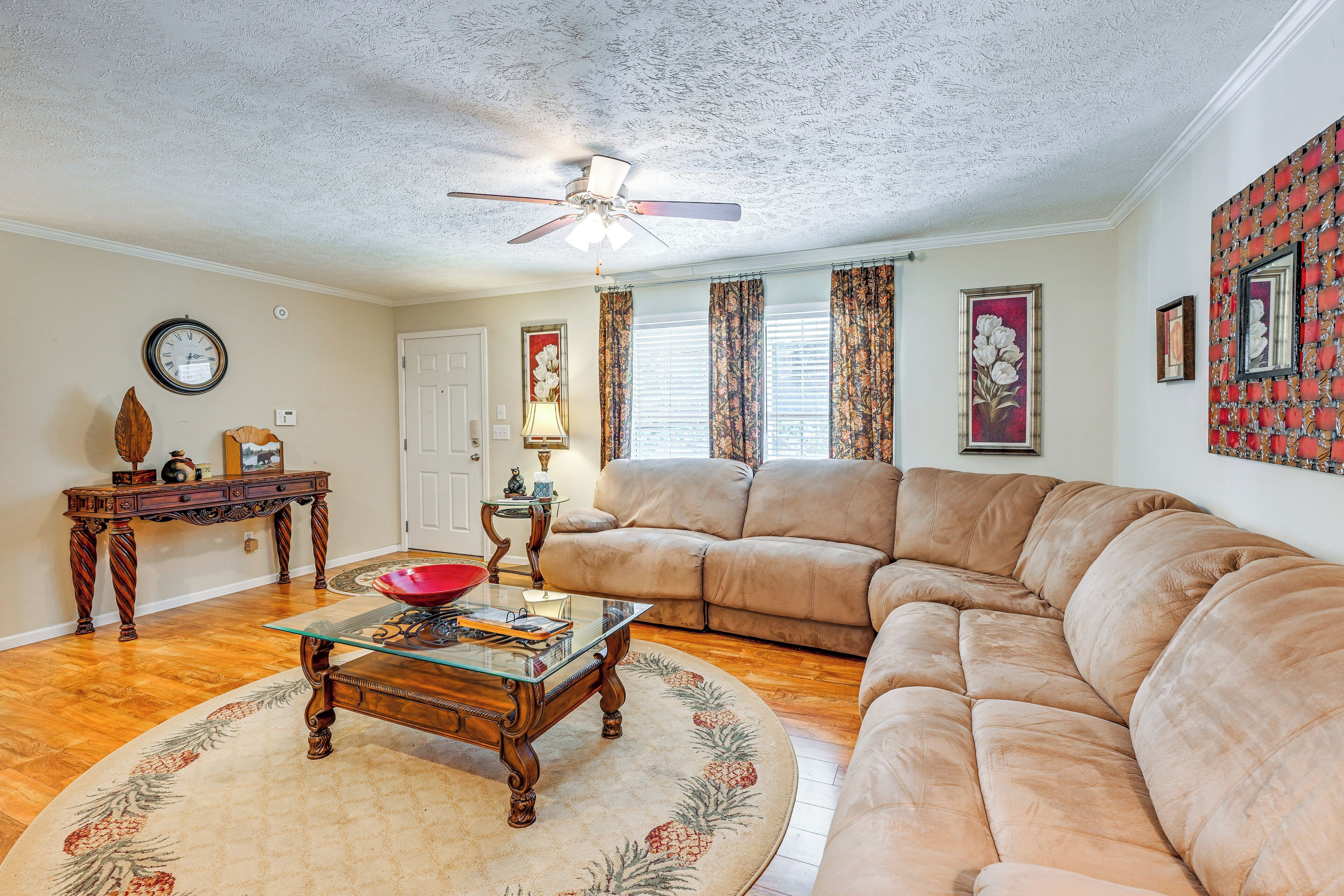 A living room with a beige sectional sofa, a glass coffee table, a wooden side table, a red bowl, a clock, and a ceiling fan.