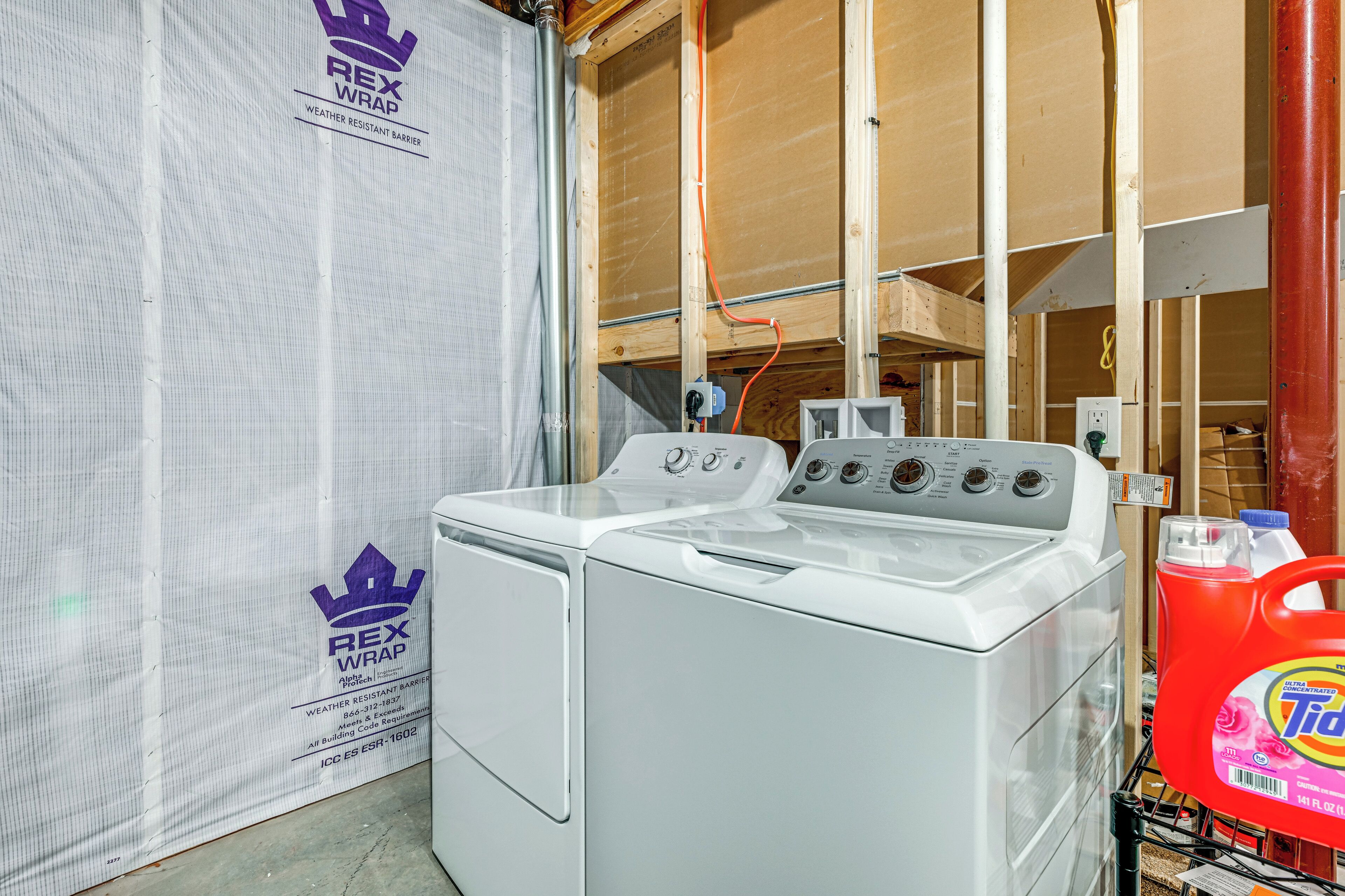 A laundry room with a washing machine and dryer, a Rex Wrap weather-resistant barrier, and a red container of Tide detergent.