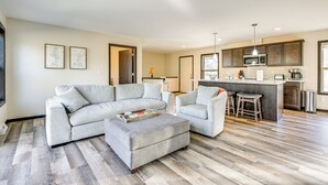 A modern living room with a grey sectional sofa, a matching ottoman, and a kitchen area with a bar. - Golf, Hike & Boat: Home w/ Fire Pit in Friendship (Friendship)