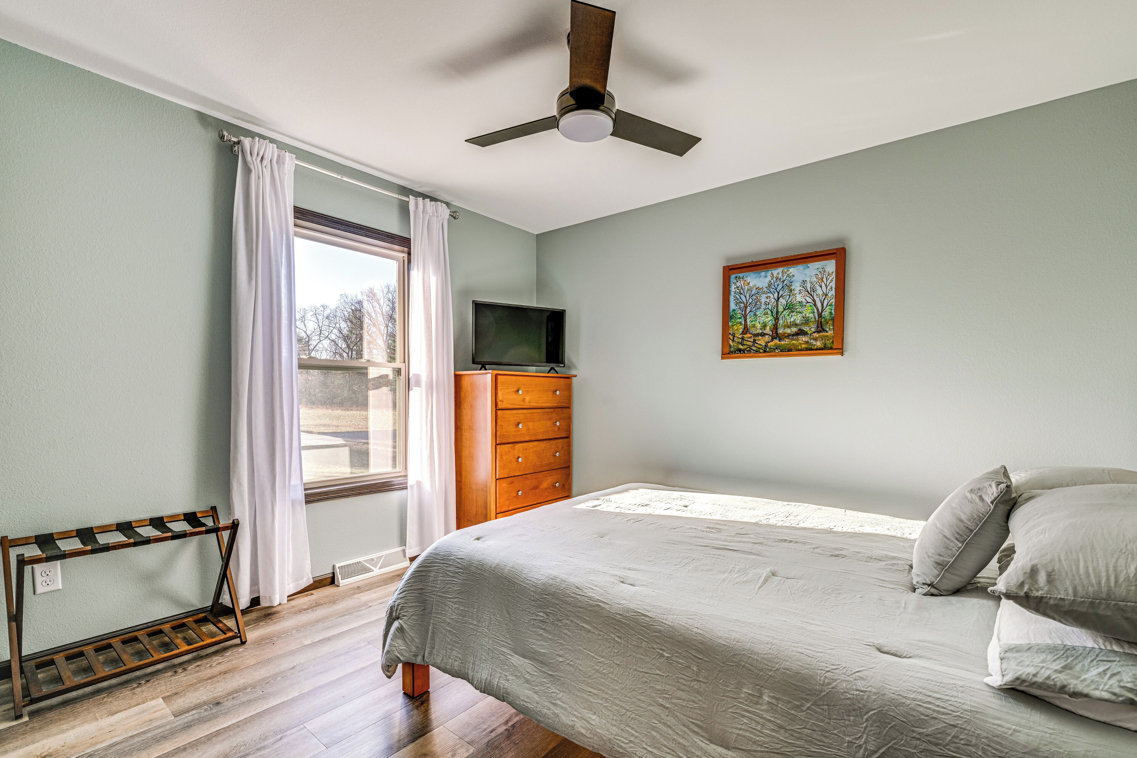 A bedroom with a bed, a dresser, a television, a painting, and a ceiling fan.