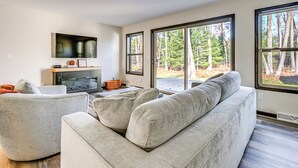 A modern living room with a large gray sectional sofa, a flat-screen TV mounted on the wall, and large windows offering a view of a forested area. - Golf, Hike & Boat: Home w/ Fire Pit in Friendship (Friendship)