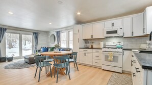 A modern kitchen with white cabinets, a central island, and a dining area with blue chairs. - 22 Mi to Boyne Mountain: Home w/ Fire Pit! (Petoskey)
