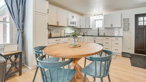 A modern kitchen with white cabinets, a wooden dining table with blue chairs, and a window with curtains. - 22 Mi to Boyne Mountain: Home w/ Fire Pit! (Petoskey)