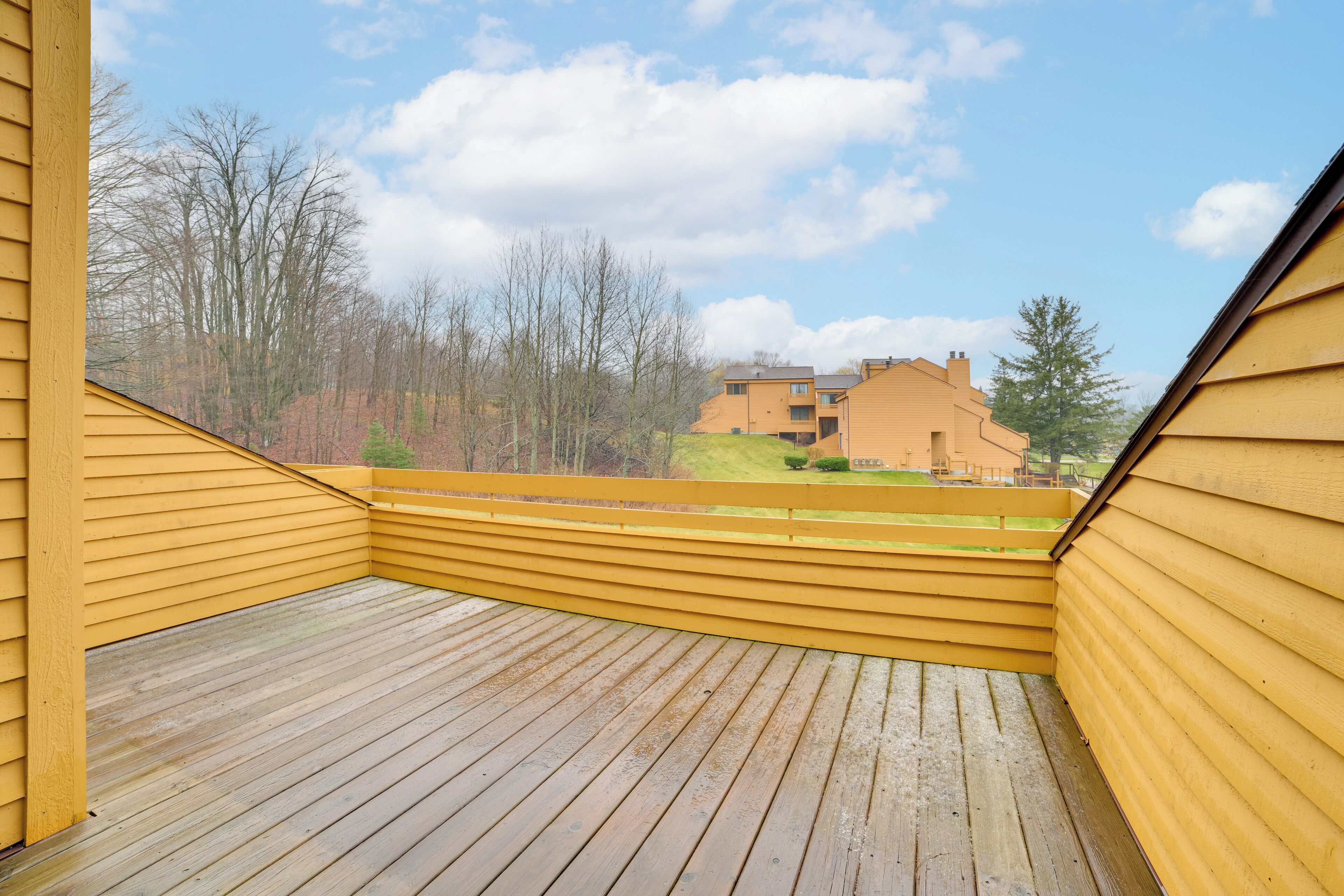 A wooden deck with yellow siding, overlooking a residential area with houses and trees.
