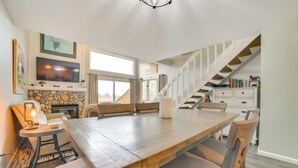 Un vaste salon doté d’une grande table en bois, de sièges confortables, d’une cheminée en pierre, d’un téléviseur à écran plat et d’un escalier menant à un niveau supérieur.