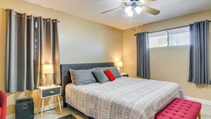 Un dormitorio con cama, mesitas de noche, una silla roja, un ventilador de techo y una ventana con cortinas.