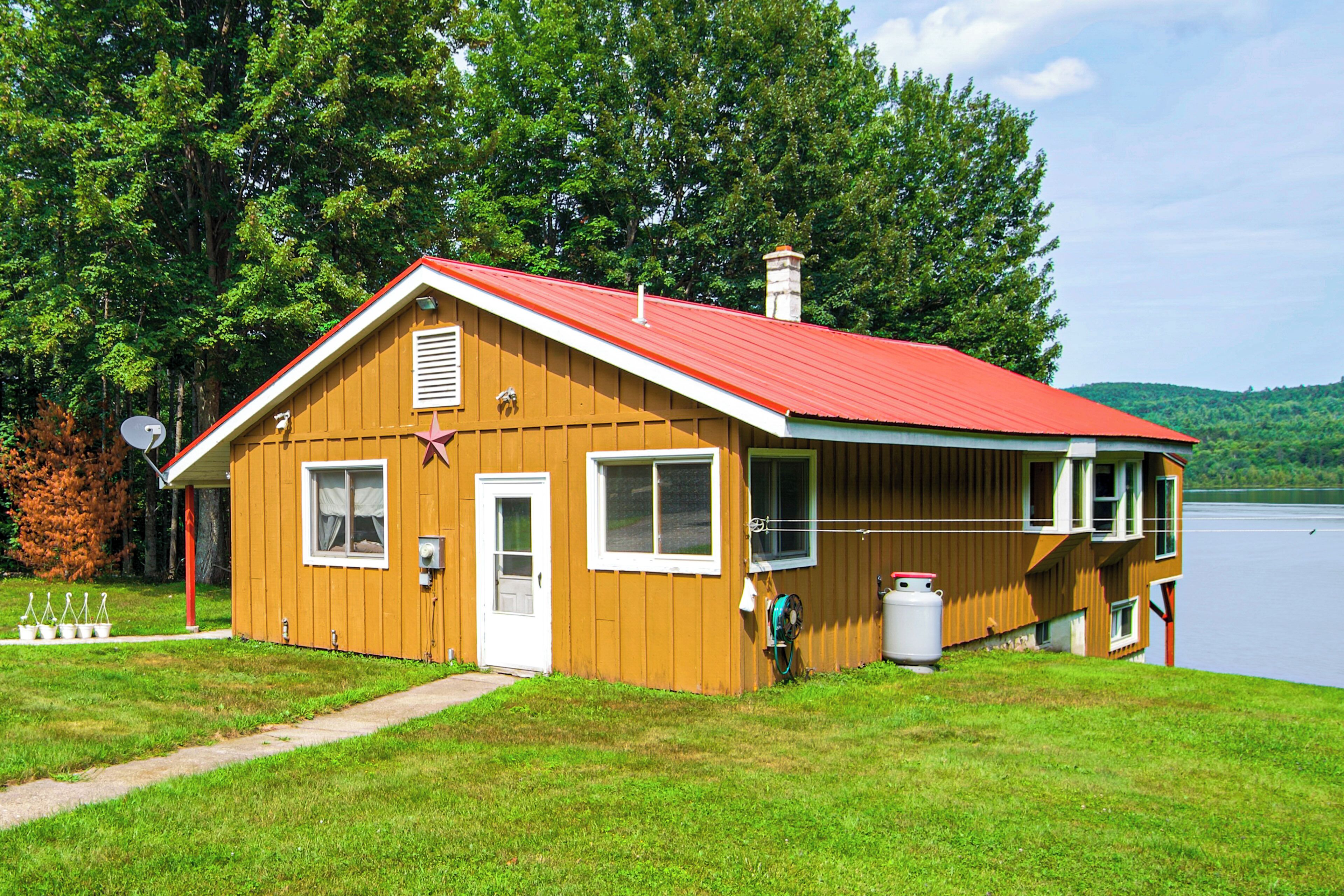 A wooden cabin with a red roof, a door, and a window, situated next to a body of water with trees in the background.