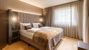 A modern bedroom with a large bed, a headboard with built-in lighting, a bedside table, and a view of the outdoors through sheer curtains. - Superior 1-bedroom Apartment (Schruns)