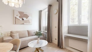 Een moderne woonkamer met een beige bank, een ronde witte tafel met gouden poten, een bankje met gekleurde kussens en een groot raam met gordijnen.