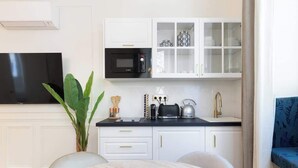 A modern kitchen with white cabinetry, a black microwave, a kettle, and a coffee maker. There is a large plant on the left and a blue upholstered chair to the right. - A Sublime 1-BR with Services in Batignolles (Paris)