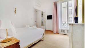 A hotel room with a bed, bedside table, lamp, and a view of the city through large windows.