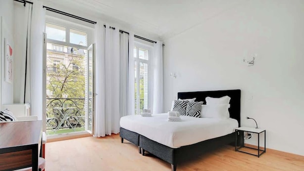 A bedroom with a bed, a bedside table, a chair, a desk, and a view of the outside through a window.