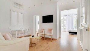 A spacious living room with a white couch, a TV mounted on the wall, and a glass table.