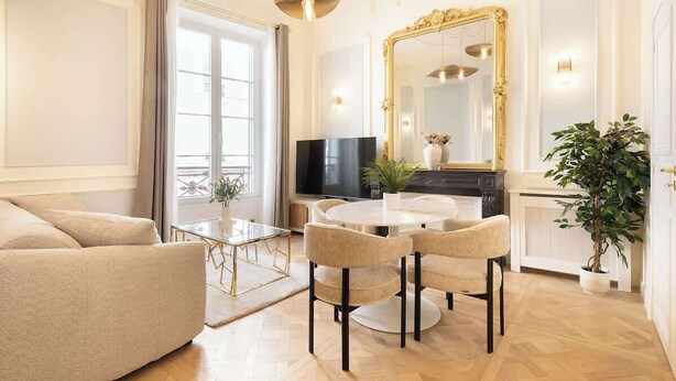 A well-lit living room with a sofa, a round dining table with chairs, a television, a large mirror, and a potted plant.