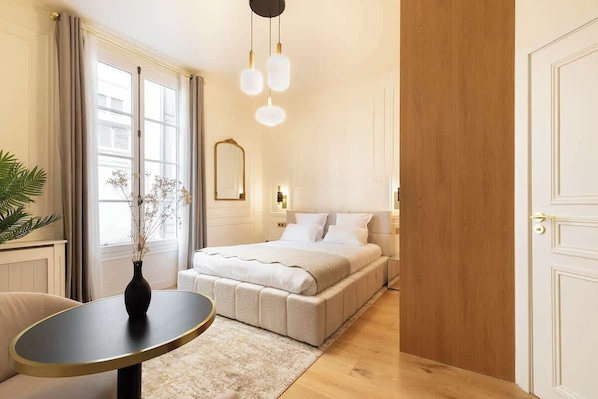 A bedroom with a bed, bedside table, and a mirror. There is a window with curtains, a plant, and a vase on the table. - A Romantic Studio with Services in Opera (Paris)