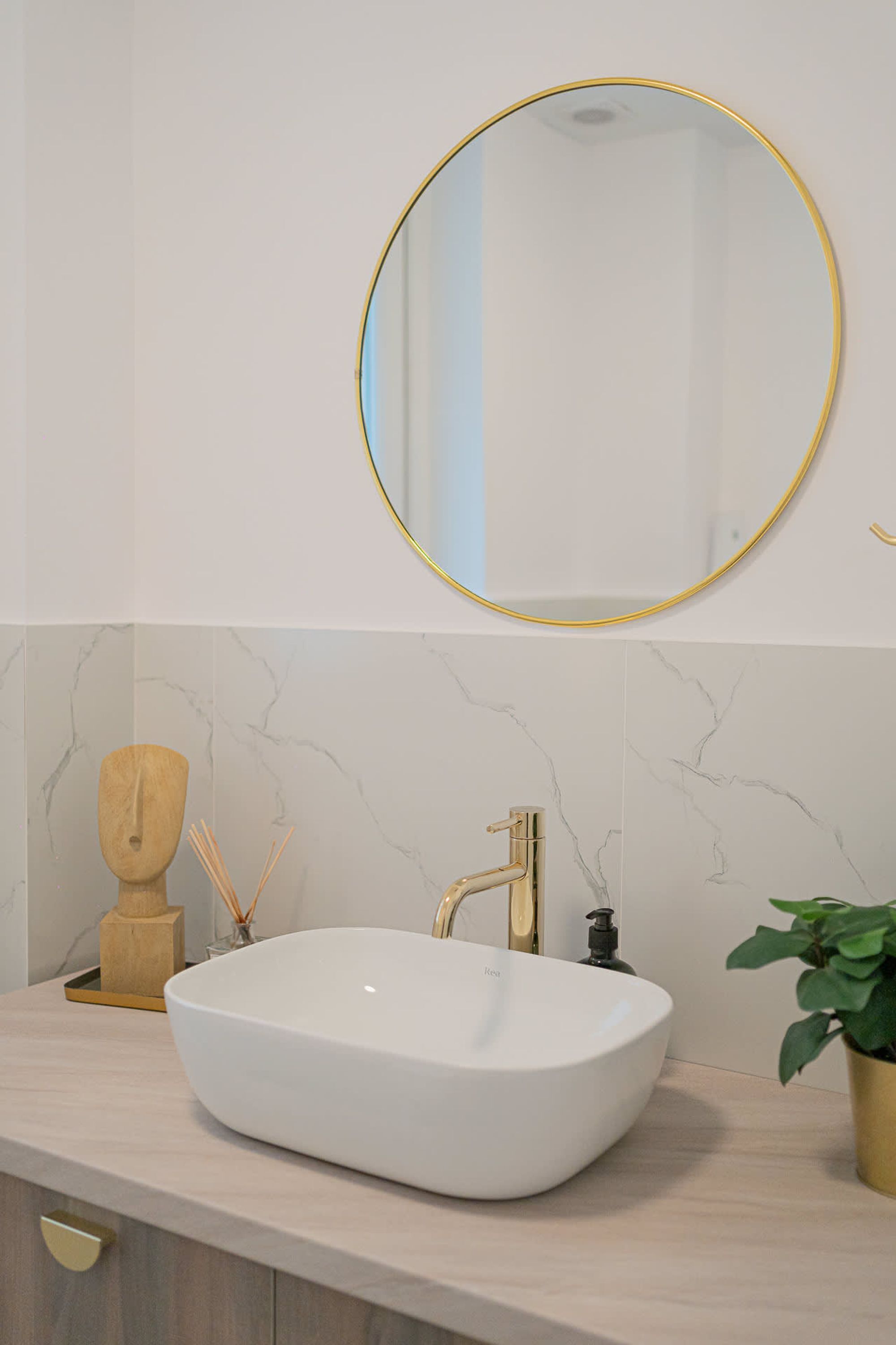 A bathroom with a white vessel sink, gold faucets, a round mirror with a gold frame, and a marble backsplash.