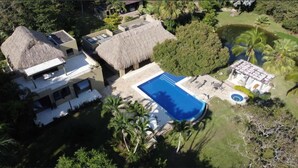 Una casa grande con piscina, construcciones de techo de paja y abundante vegetación.