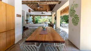 Una mesa de comedor de madera con sillas blancas de mimbre, un ventilador de techo y vistas a un jardín exuberante a través de un patio abierto.