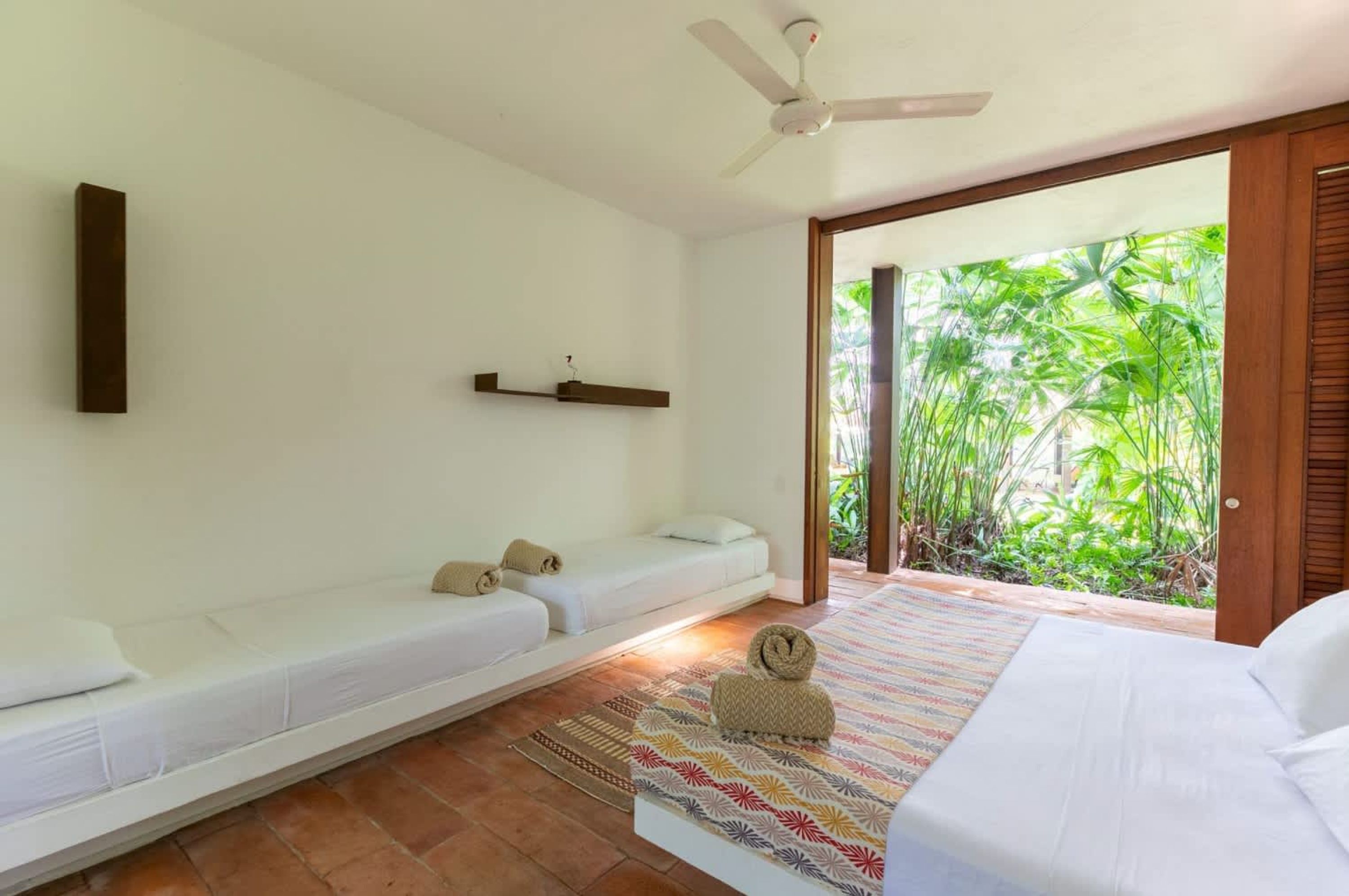 A room with a bed, a sofa, a ceiling fan, and a view of greenery outside.