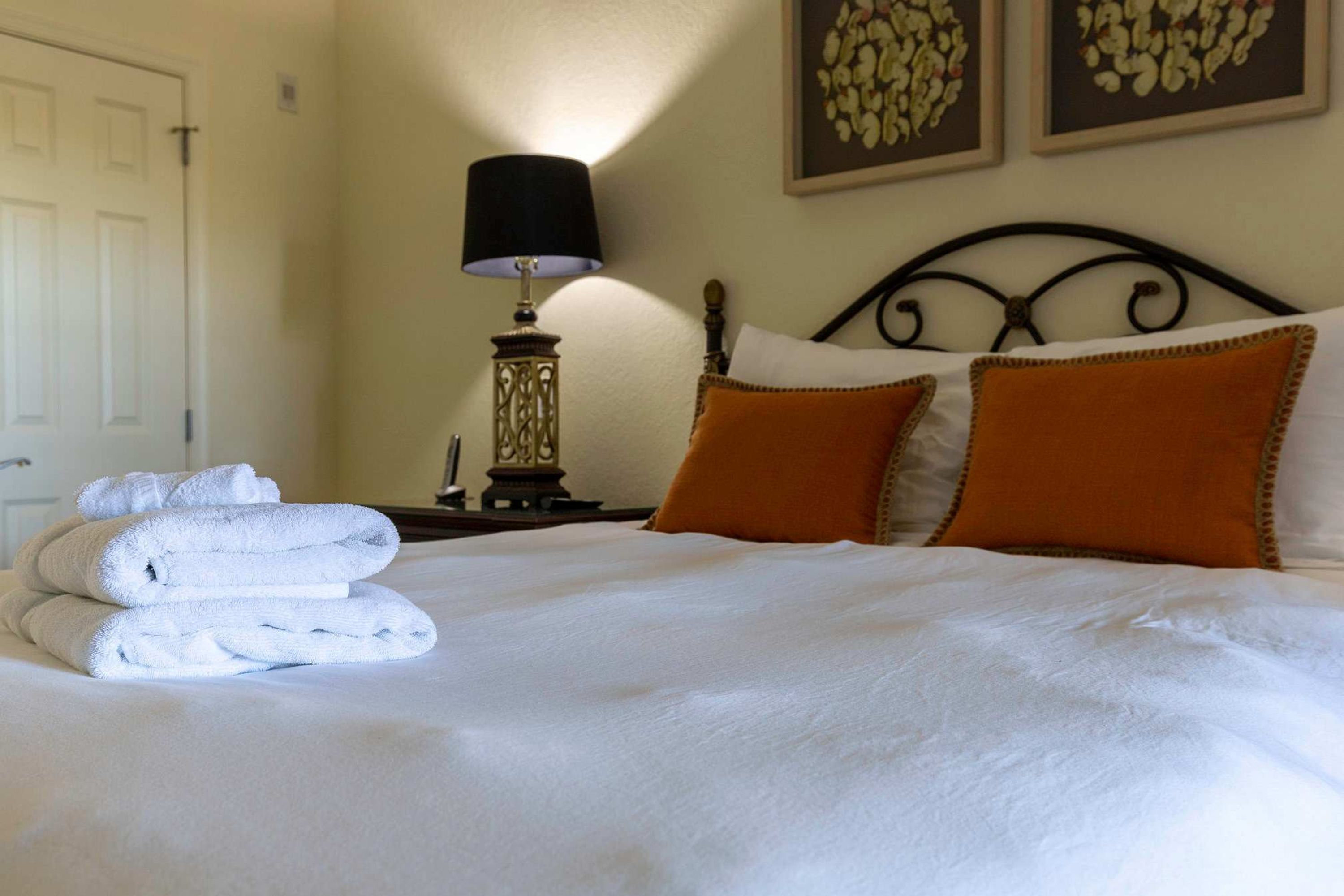 A neatly made bed with white linens and two orange pillows, a bedside lamp, and a wall clock.