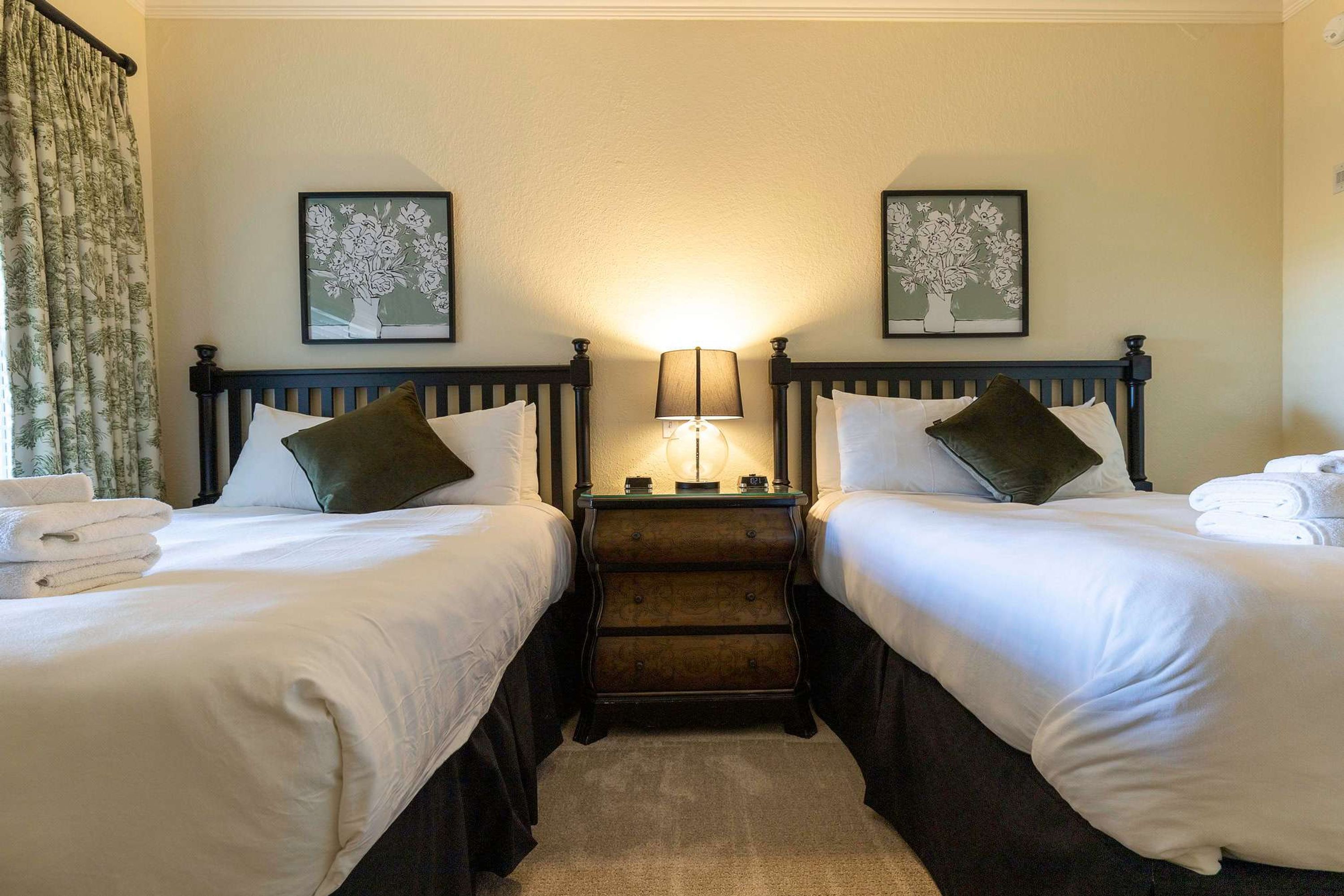 A hotel room with two single beds, a nightstand with a lamp, and two framed floral artworks on the wall.