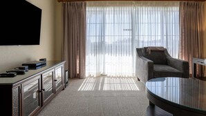 Habitación de hotel con televisor de pantalla plana montado en la pared, una mesa de centro de vidrio, un sofá y un ventanal con cortinas translúcidas.