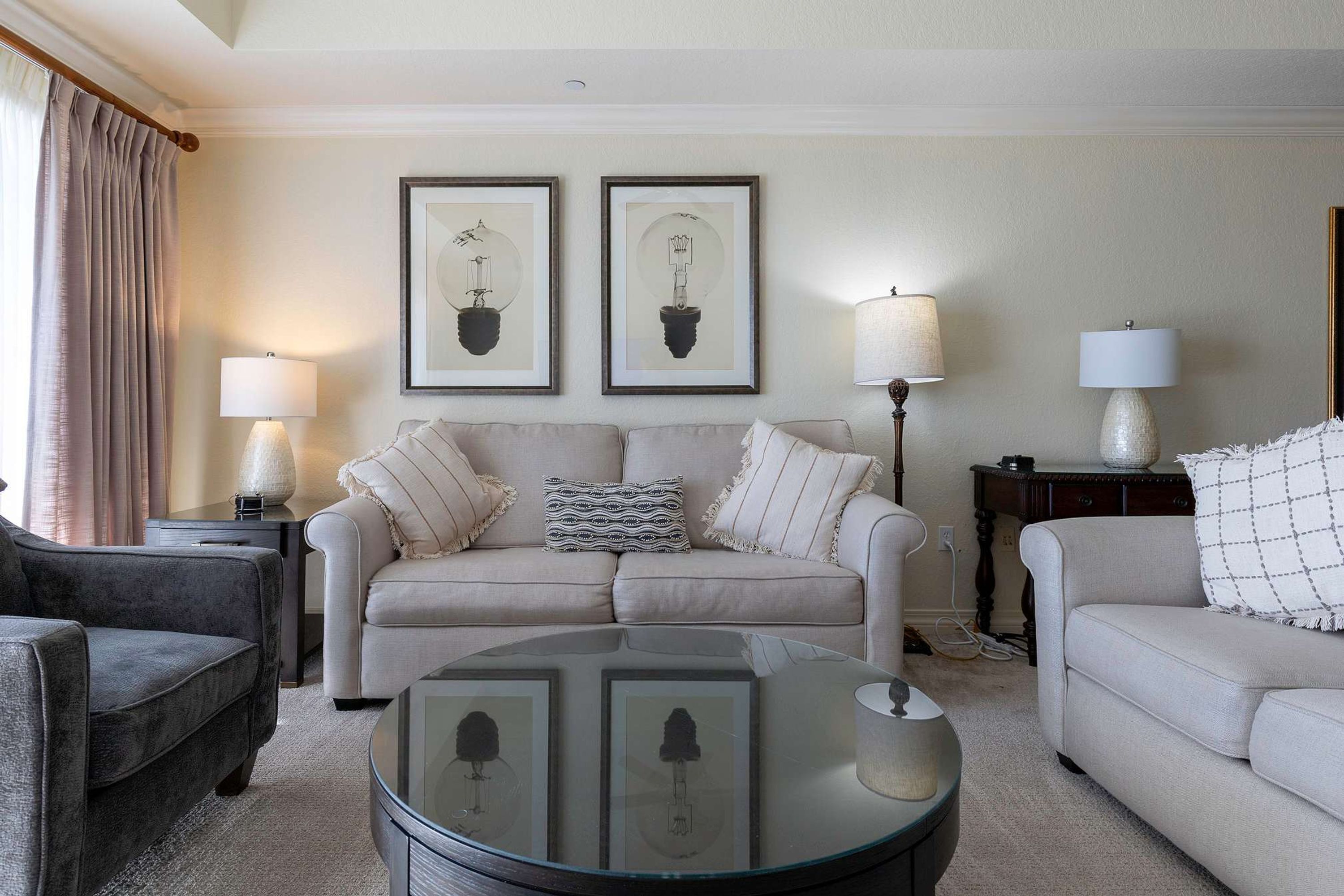 Una sala de estar con un sofá color beige, un sillón oscuro y una mesa de centro de vidrio. Hay dos cuadros en la pared y dos lámparas sobre mesitas de noche.