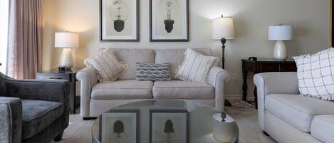 A living room with a beige sofa, a dark armchair, and a glass coffee table. There are two framed pictures on the wall and two lamps on side tables.